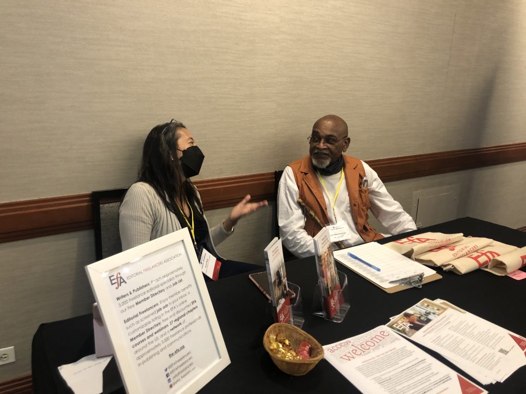 Two people in conversation behind a table covered with marketing materials.