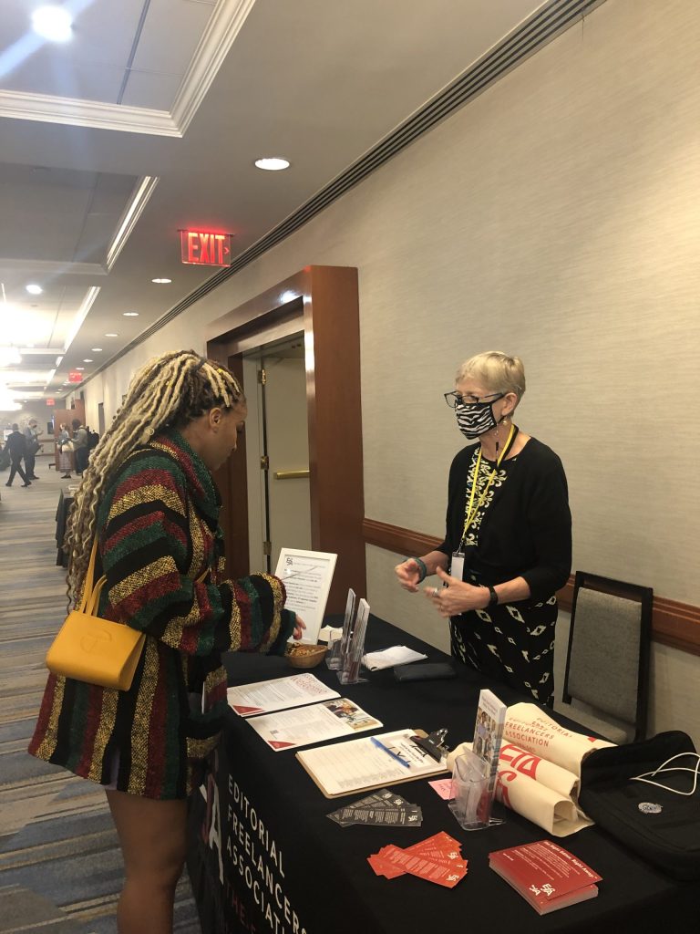 Two people in conversation on opposite sides of a table covered with marketing materials.