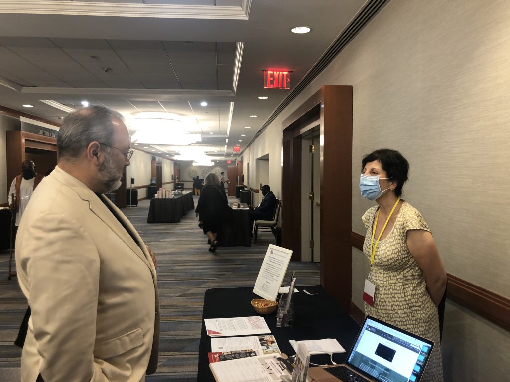 A caucasian man and woman standing in conversation on opposite sides of a table covered with flyers, brochures, and other promotional items.