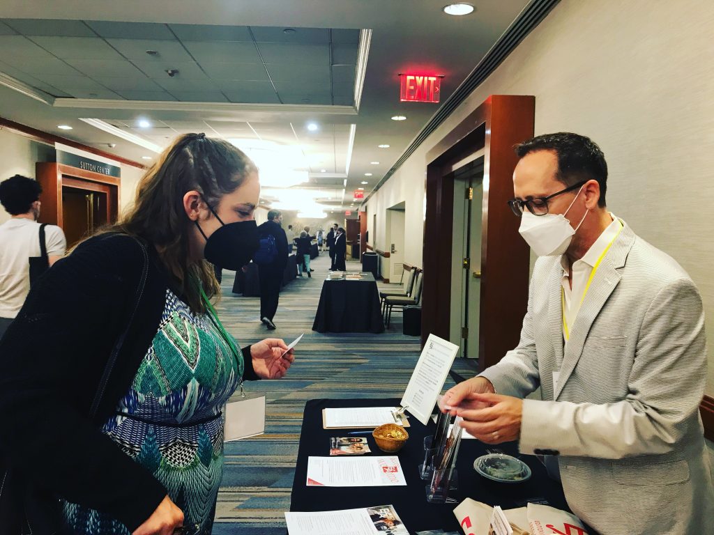 Two people in conversation on opposite sides of a table covered with marketing materials.