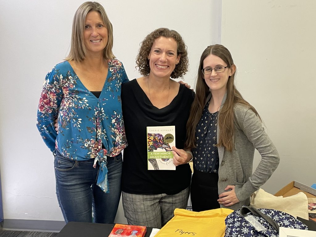 Three women standing behind a table; the one in the middle holding up a book