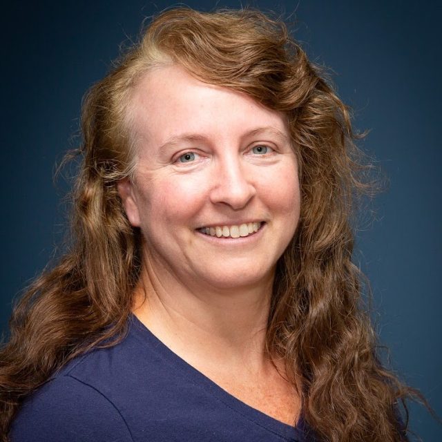 Headshot of Cyndi Sandusky with long red hair in a blue top in front of a blue gradient background