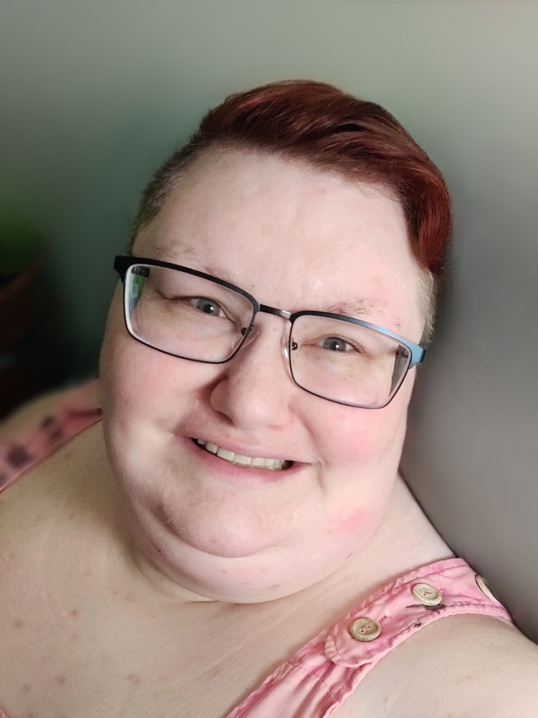 Headshot of Dayna Reidenouer with cropped red hair, wearing black eyeglasses and a pink sleeveless top in front of a gray gradient background