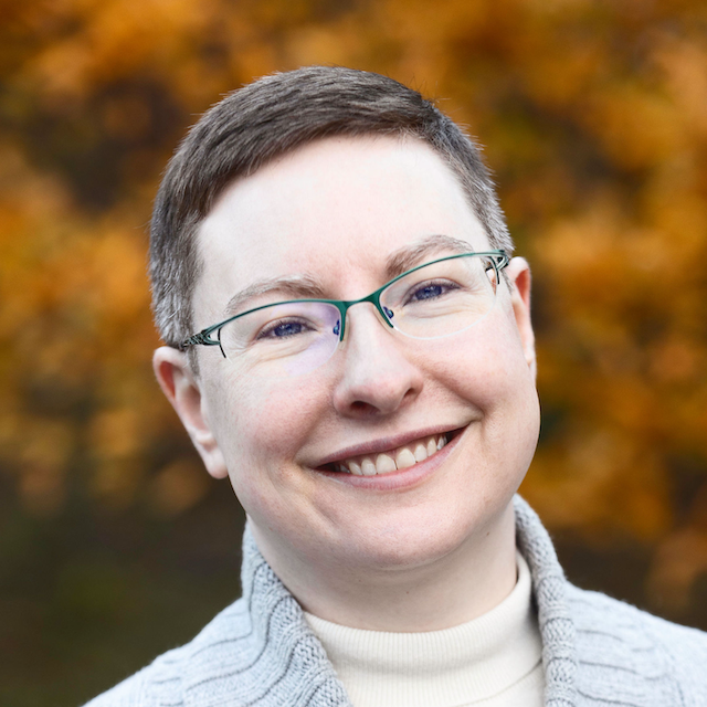 Smiling person with glasses dressed in a white turtleneck under a gray sweater