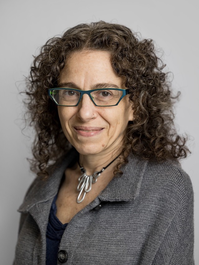 Smiling woman with glasses and necklace, dressed in a black top under a gray jacket