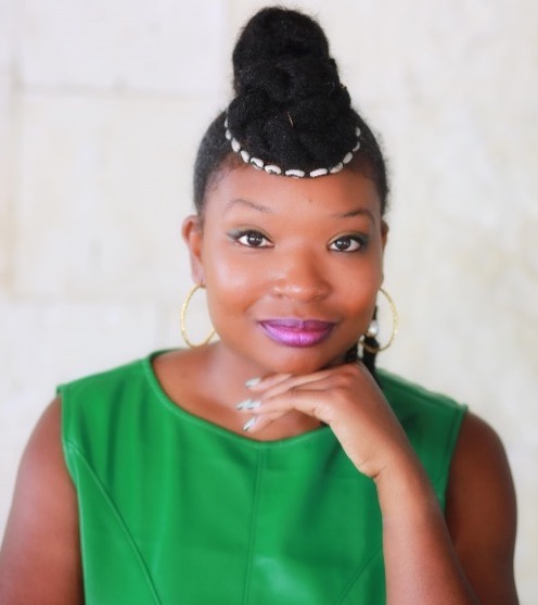 Smiling Black woman wearing a green sleeveless top, gold earrings, and a white headband, propping up her chin with with her hand.