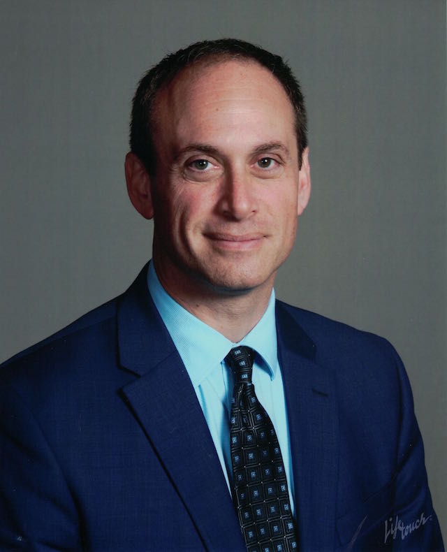 Smiling man with a light blue shirt and dark blue tie under a dark blue jacket